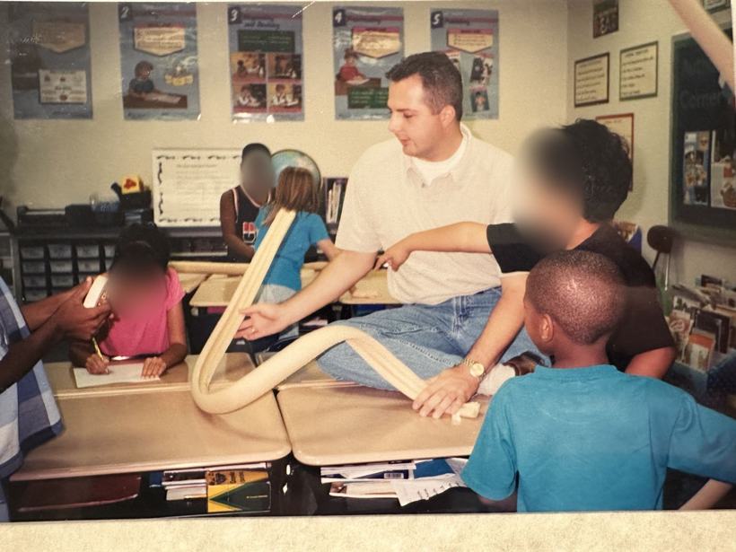 Shane as a young teacher on a desk with second graders and a foam roller coaster track. Student faces blurred.