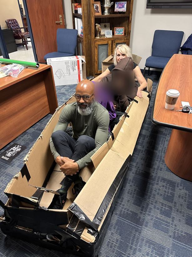 Adults and students sitting in a full-size duct-taped cardboard boat. Student faces blurred.